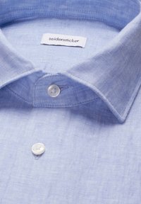 Chemise bleu clair à boutons avec col pointu et boutons blancs, présentant une étiquette de marque "seidensticker" visible à l'intérieur du col.