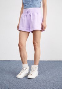 Person som bär ljuslila drarem shorts, vita högklackade sneakers och en ärmlös kort denimjacka, står på en blå matta.