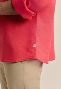 Camisa de lino roja con mangas enrolladas, presenta un borde rematado y un pequeño detalle bordado en azul y blanco cerca del fondo.