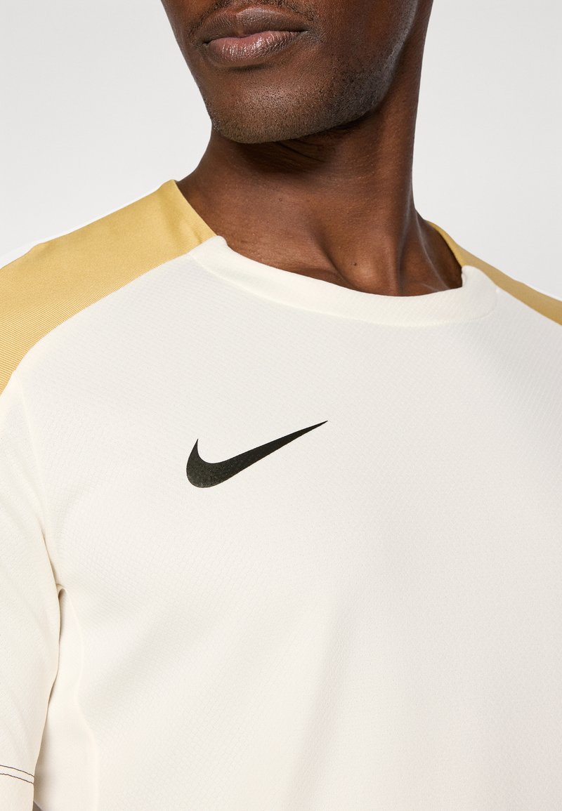 T-shirt de sport Nike crème avec épaules dorées, arborant un logo noir sur la poitrine, fabriqué en tissu texturé pour le confort et la performance.