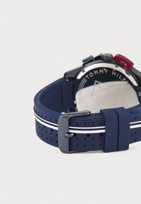 Cinturino per orologio in silicone blu navy con design perforato e strisce bianche, dotato di hardware in argento e un accento rosso sul pulsante di attivazione.