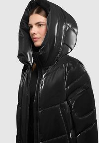 Sort, blank pufferjakke med stor hætte, kwartet design, frontlynlås og sidelommer. Synlige syninger og lynlåslomme på ærmet.