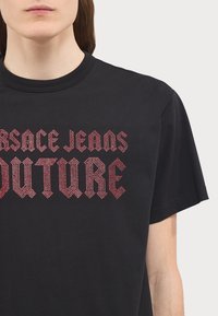 Černé bavlněné tričko s výrazným červeným textem z kamínků "VERSACE JEANS FUTURE", s kulatým výstřihem a krátkými rukávy.