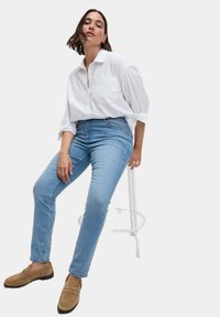 Bílá košile s dlouhými rukávy, světle modré džínové kalhoty s vysokým pasem a béžové loafers; sedící na bílém stoličce na neutrálním pozadí.