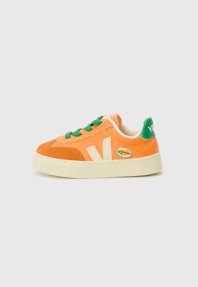 Sneaker arancioni con lacci verdi, con una parte superiore in suede e tela. Suola in gomma bianca e dettaglio circolare del logo sul lato.