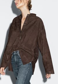 Bruine suède oversized shirt met een notch kraag, vooraan met ritssluiting en zichtbare stikdetails, gecombineerd met blauwe high-waisted jeans.