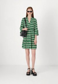 Zeleno obarvana obleka s trikotnimi rokavi do komolca, večstopenjski dizajn in črna torbica. Model nosi črna sončna očala in platformne sandale.