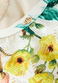 Kukkakuvioinen kangas keltaisin ja vihrein kuvioin. Pinnassa on tekstuuri ja kauluksen läheisyydessä kultainen pyöreä kiinnitin.