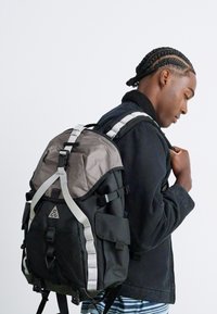 Young man with braided hair wearing a black jacket and carrying a large black and gray backpack with white straps, standing against a plain background.