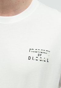 Λευκό βαμβακερό t-shirt με μαύρο εκτυπωμένο λογότυπο, όπου το "PROPERTY OF" είναι διαγραμμένο και από κάτω αναγράφεται "D.I.E.S.E.L". Ομαλή υφή, λαιμόκοψη τύπου crew.