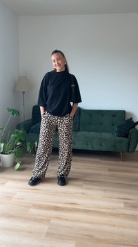 Svart oversized t-skjorte, beige leopardskinnbukser og svart fottøy. Innendørs setting med grønn sofa og potteplante. Naturlig belysning.