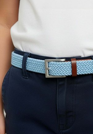 Ceinture tressée en tissu bleu clair avec boucle argentée, portée avec un pantalon bleu marine et un haut blanc rentré.
