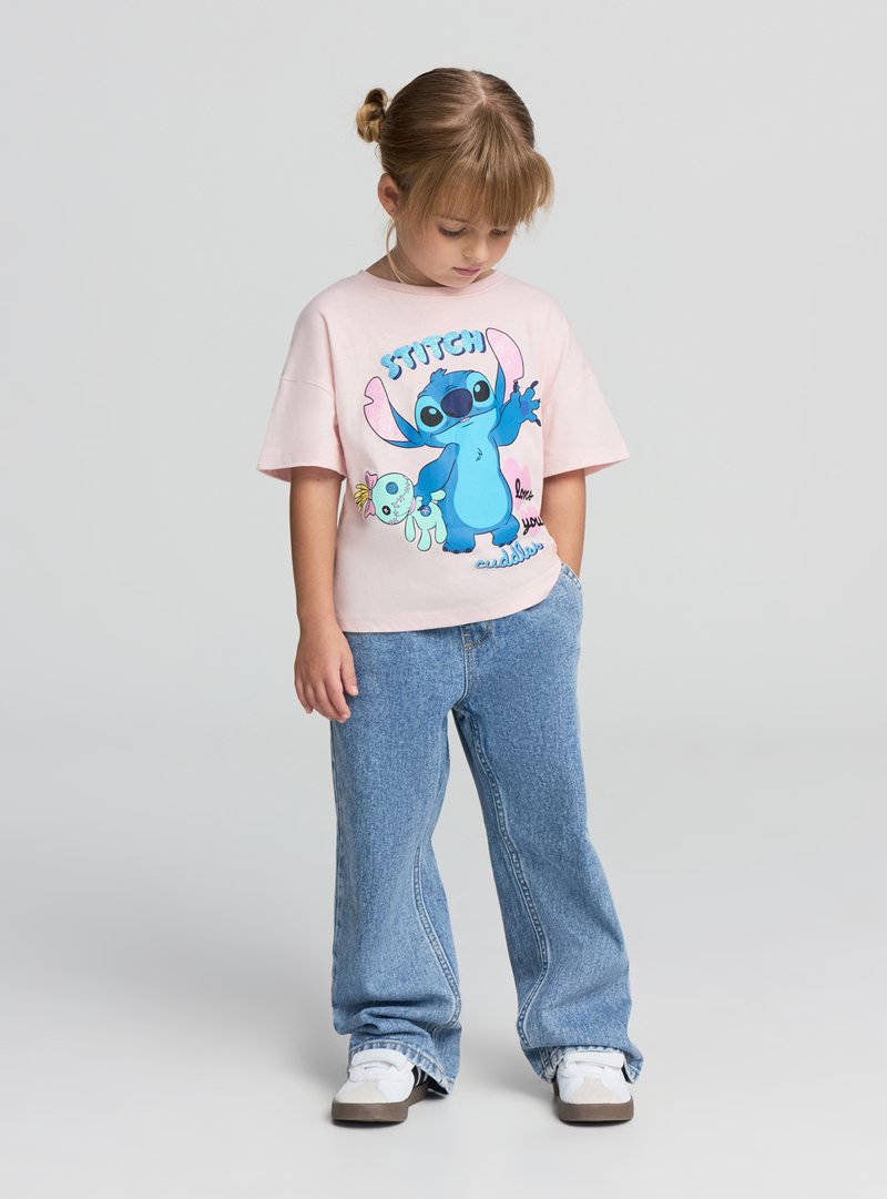 Młoda dziewczyna z blond włosami związanymi w kok, ubrana w różowy t-shirt z grafiką Stich, niebieskie dżinsy i białe trampki, stojąca z rękami w kieszeniach.