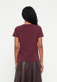 Persoon met lang zwart haar, gekleed in een kort mouw maroon shirt en een donkerbruine leren rok, staand met het gezicht naar een effen witte achtergrond.