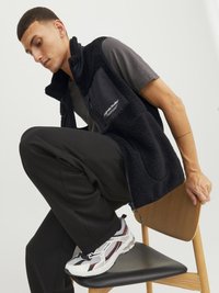 Chaleco de forro polar negro con un bolsillo con cremallera, usado sobre una camiseta gris, combinado con pantalones negros y zapatillas blancas con detalles en rojo.