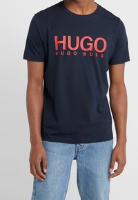 Námořnická modrá tričko s krátkým rukávem, na kterém je velký červený nápis "HUGO" a pod ním menší nápis "HUGO BOSS". Vyrobeno z bavlny se standardním střihem.