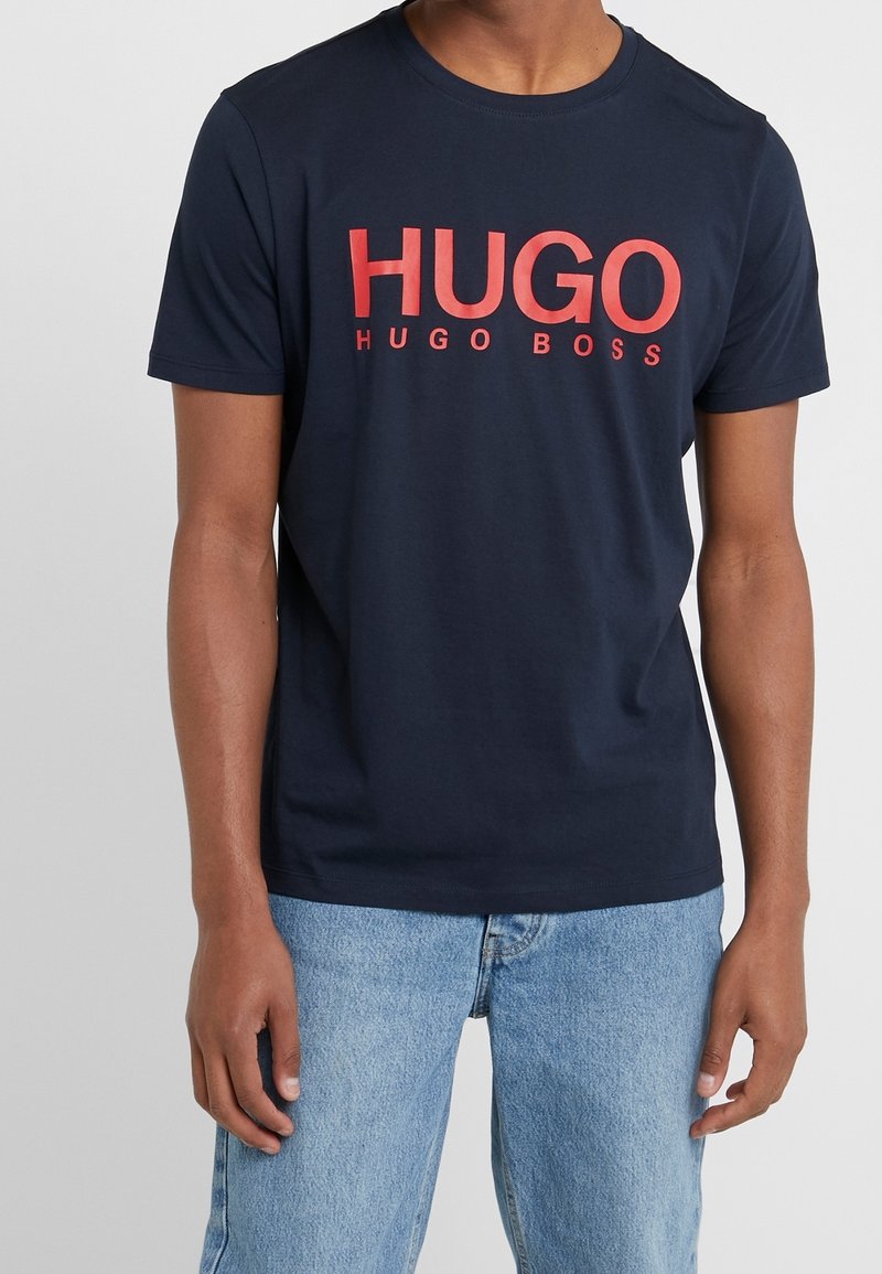 Námořnická modrá tričko s krátkým rukávem, na kterém je velký �červený nápis "HUGO" a pod ním menší nápis "HUGO BOSS". Vyrobeno z bavlny se standardním střihem.