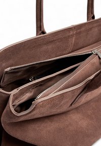 Brun mockaskinn toteväska med två handtag, som har ett fodrat fack och en öppen ficka inuti. Mjuk textur med sömnadsdetaljer.