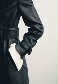 Cappotto in pelle nera con vita regolabile e maniche larghi. Tessuto bianco sbircia dal polsino. Texture liscia e design su misura.