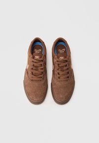 Brown suede sneakers with a rounded toe, flat laces, and a blue inner lining. Features a gold Nike swoosh logo on each side. Rubber sole.