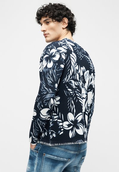 Homme aux cheveux bouclés portant un pull marine avec de grands motifs floraux blancs, associé à un jean bleu, tourné légèrement de côté.