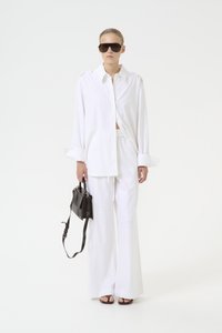 Vit oversized skjorta och vida byxor i lätt tyg. Svart handväska med rem. Modell som bär stora mörka solglasögon och sandaler.