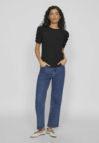 Čierny pletený top s textúrou a balónovými rukávmi, kombinovaný s džínsami s rovnými nohavicami v modrej farbe a svetlými plochými topánkami. Modelka stojí s rukami na bokoch.