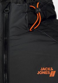 Detale czarnej kurtki z zamkiem błyskawicznym, pomarańczowym ściągaczem oraz pomarańczowym logo "JACK & JONES" na tkaninie.
