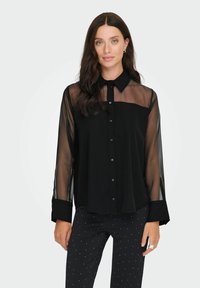 Crna košulja dugih rukava s prozirnim rukavima, čvrstim prednjim dijelom i klasičnom kragnom. Materijal izgleda glatko s blagim sjajem.