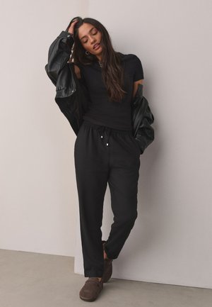 Femme appuyée contre un mur blanc, portant un haut noir à manches courtes, un pantalon noir, en train d’enlever partiellement une veste noire, et des chaussures marron faciles à enfiler.