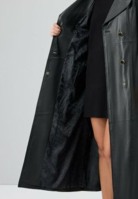 Lang zwart leren jas met een dubbelbreasted ontwerp. Het interieur heeft een glad zwarte voering met cirkelpatronen. Functionele knopen zijn zichtbaar.