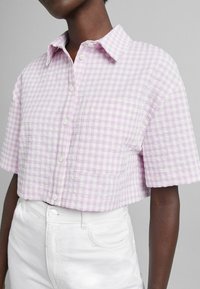 Kortärmad, kragad blus i lavendel- och vit rutigt mönster. Strukturerad tyg med en ficka på vänster sida, knäppning fram.