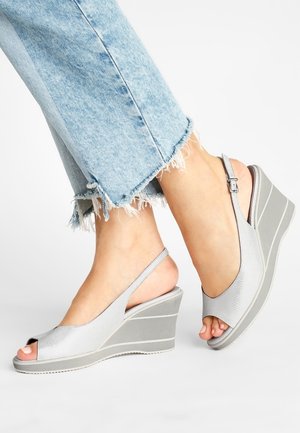 Füße tragen hellgraue Keilsandalen mit Peeptoe und Knöchelriemen, kombiniert mit hellblauen Jeans mit ausgefranstem Saum und verkürzter Länge.