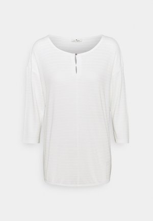 Blouse blanche à manches longues avec un col rond et un petit détail d’ouverture boutonnée à l’avant sur fond uni.