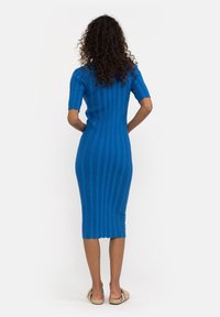 Abito a maglia di media lunghezza in blu brillante, con texture a coste verticali, maniche corte e orlo ondulato. Indossato con scarpe flat di colore chiaro.
