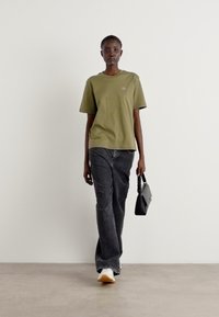 Olivgrünes Baumwoll-T-Shirt, lockere Passform, kombiniert mit dunklen Denim-Jeans. Das Modell trägt eine kleine schwarze Handtasche und weiße Sneakers.