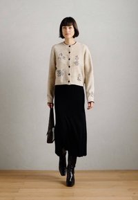 Μπεζ πλεκτό ζακέτα με βοτανική κεντημένη διακόσμηση, με κουμπιά μπροστά, συνδυασμένη με μακριά μαύρη ριγέ φόρμα και μαύρες μπότες μέχρι το γόνατο.