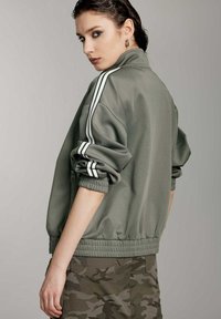 Chaqueta bomber verde oliva con rayas blancas en las mangas, puños y cinturilla acanalados. Tejido suave y liso. Combinada con pantalones cortos de camuflaje.