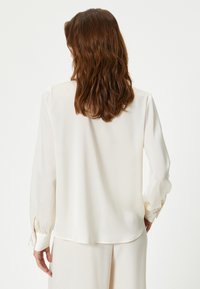 Creme satinbluse med lange ærmer, der har knaplukning ved manchetterne og en glat overflade. Bagstykket er en smule længere end forsiden.