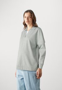 Top de manga larga gris claro con cuello, que presenta un corte amplio y una textura sutil. Combinado con pantalones de mezclilla azul claro.