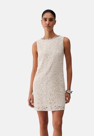 Donna che indossa un mini abito smanicato color beige in pizzo con motivi floreali e un braccialetto metallico, in piedi contro uno sfondo chiaro e uniforme.