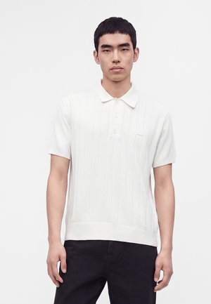 Jeune homme portant un pull polo blanc à manches courtes et un pantalon noir, debout devant un fond blanc uni.