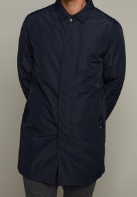 Abrigo ligero en azul marino, resistente al agua, con cuello puntiagudo, cierre frontal de botones y dos bolsillos laterales. Textura suave, diseño hasta la rodilla.