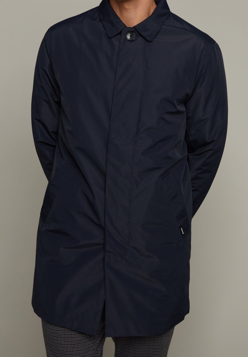 Abrigo ligero en azul marino, resistente al agua, con cuello puntiagudo, cierre frontal de botones y dos bolsillos laterales. Textura suave, diseño hasta la rodilla.