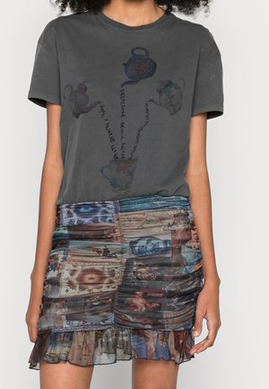 Kvinde iført mørkegrå T-shirt med grafik af tekander, der hælder i en kop, og en multifarvet rynket mini nederdel med lapværksprint.