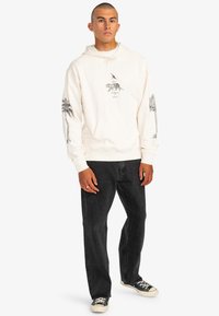 Light cream hoodie with palm tree and tiger graphic prints, soft texture, drawstring hood, front pocket, paired with black jeans and canvas shoes.