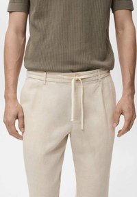 Beige linnen broek met een trekkoord in de taille, met een gladde textuur en plooidetails. Gemaakt met een gestructureerde olijfgroene gebreide top.