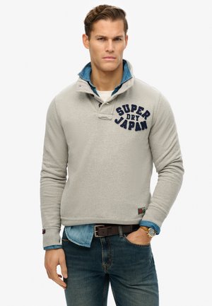 Sudadera de forro polar gris con el bordado en azul marino "SUPER DRY JAPAN" en el pecho, superpuesta a una camisa de mezclilla, con un cuello alto y un detalle de botones.