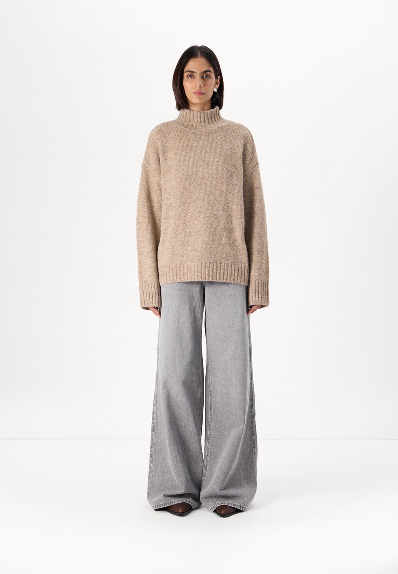 Beige knitted turtleneck sweater with wide sleeves, paired with light gray wide-leg jeans and pointed-toe black shoes.