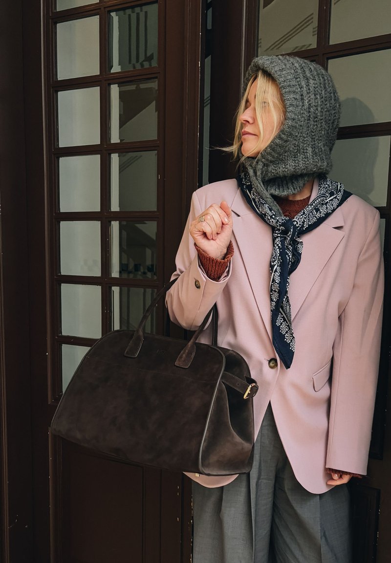 Bruine suède handtas met dubbele handvatten, gecombineerd met een lichtroze blazer over een roestkleurige trui en een blauw patroon sjaal.